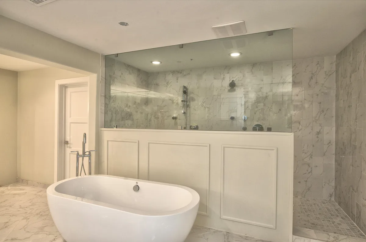 A luxurious ensuite bathroom with a freestanding soaking bathtub, a glass-enclosed shower, and marble walls. The room is bathed in soft, natural light, evoking a spa-like atmosphere.