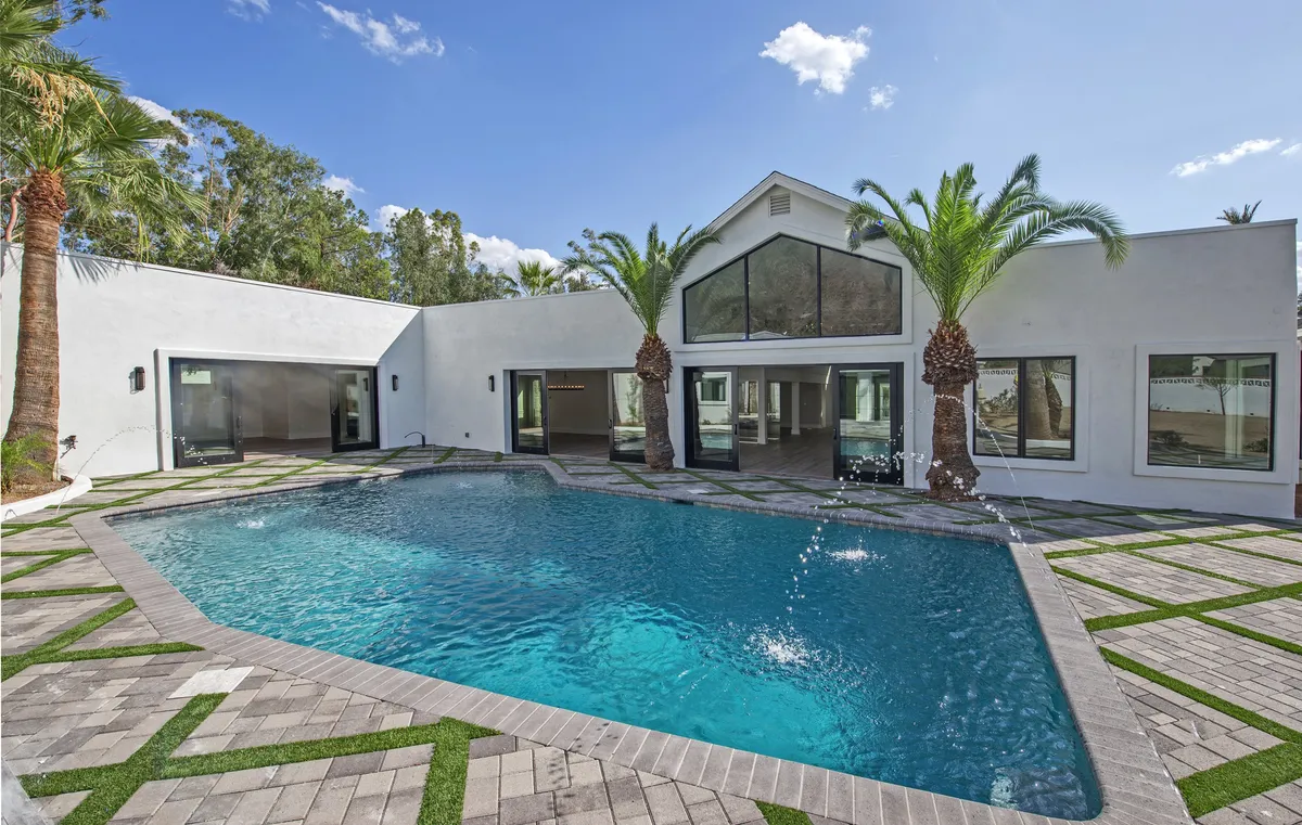 A luxurious, resort-style backyard featuring a sparkling swimming pool surrounded by palm trees, large windows connecting indoor living spaces, and landscaped pathways. The scene captures the transition from the indoors to an inviting outdoor oasis, with hints of sunset lighting adding to the ambiance.