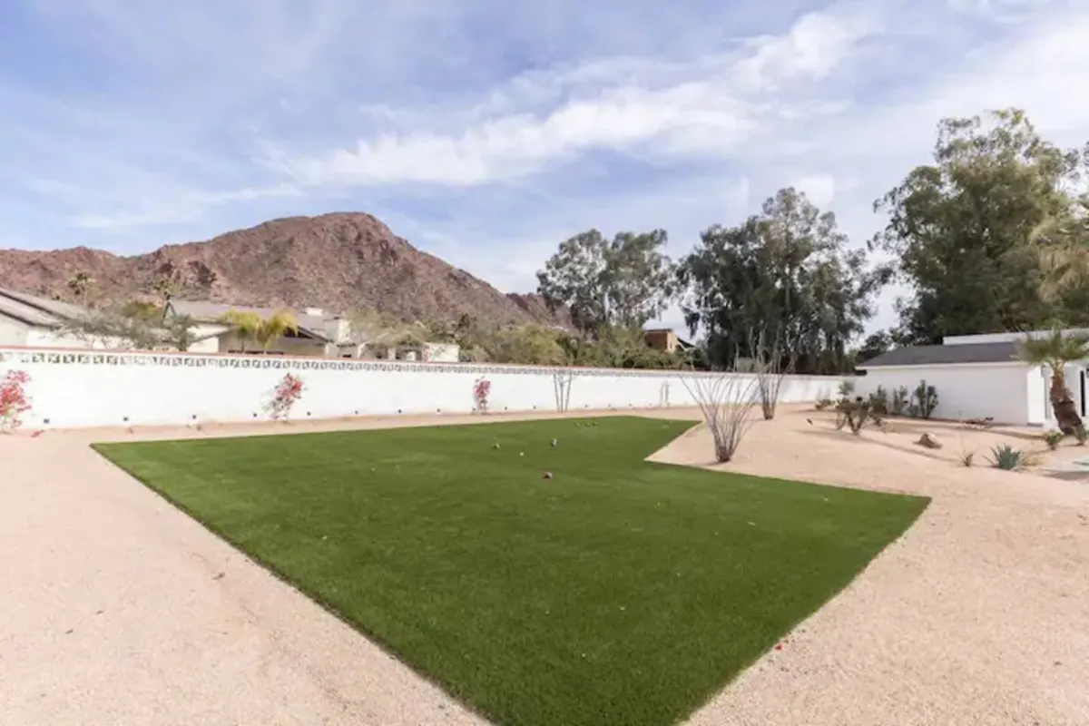 A serene exterior scene featuring a lush green lawn, desert landscaping, a white wall, and Camelback Mountain in the distance. The open space is perfect for gatherings or relaxation, with the golden hour lighting creating a welcoming, luxurious feel.