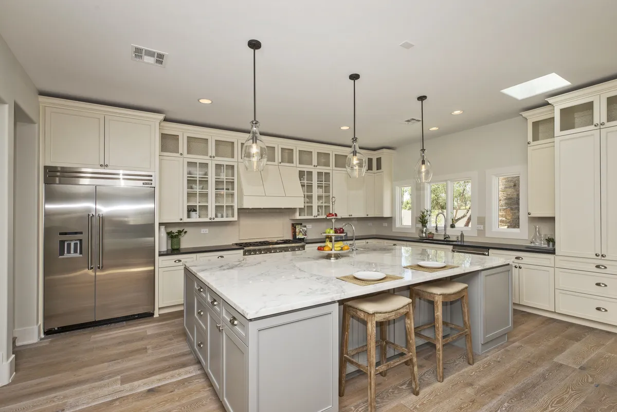 This image showcases a modern kitchen with a large island, stainless steel appliances, pendant lighting, and abundant natural light streaming through large windows. The scene suggests a space designed for both culinary activity and social interaction, with plenty of room for cooking, entertaining, and casual dining.