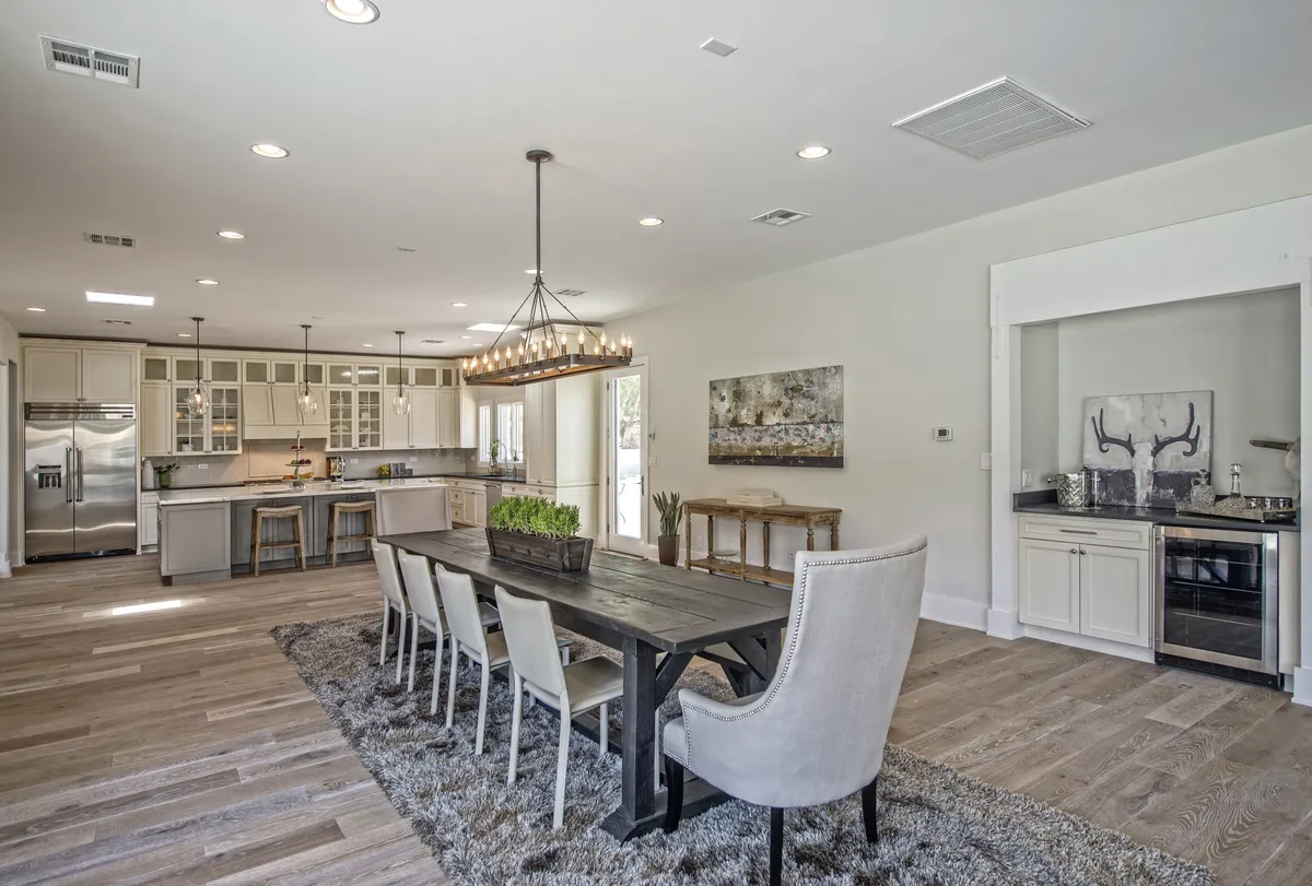 A wide-angle view that includes a long dining table under a chandelier, a kitchen island in the background, and large windows letting in natural light. The arrangement highlights both the formal dining area and its proximity to the kitchen, suggesting seamless flow for serving meals and hosting gatherings.