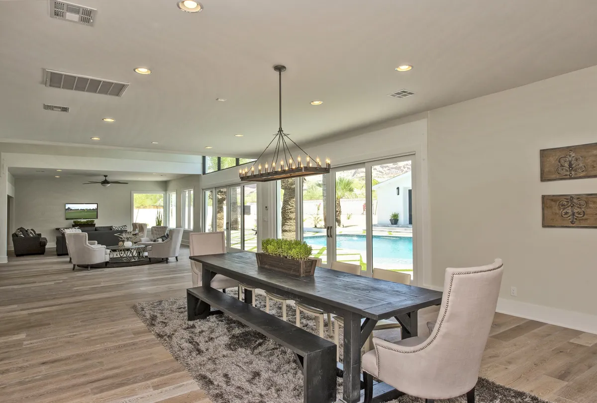 A welcoming dining area with a large table set beneath an elegant chandelier, adjacent to a comfortable seating area. Large windows offer views of the pool and allow natural light to flood the space, enhancing the ambiance for celebrations or casual relaxation.