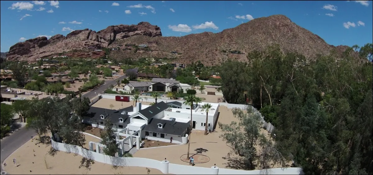 A striking view of the estate's exterior, featuring white walls set against a dramatic mountain backdrop, meticulously landscaped gardens, and a paved driveway. The landscaping is lush yet distinctly desert-inspired, and the sky hints at early morning or sunset, adding a tranquil mood.