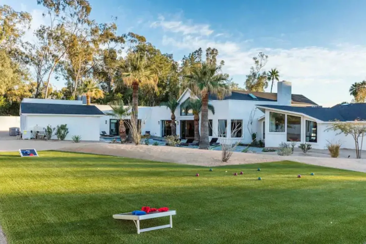 This exterior image shows a modern house with a lush lawn set up for outdoor games, surrounded by palm trees. The scene is bright and playful, suggesting a space where families can gather for fun activities and celebrations in a spacious backyard.